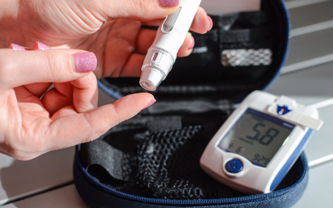 Woman with glittery pink nail polish uses a lancing device on her finger next to a digital glucometer displaying a reading of 5.8, sitting in a blue carrying case.