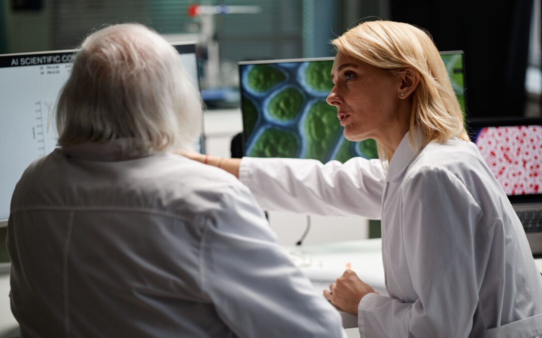 Two researchers in lab coats collaborating; a woman points to a computer screen displaying green cellular structures while advising an older man.